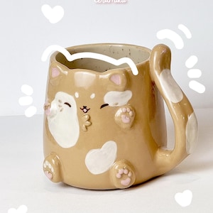 Può includere: Una tazza in ceramica color marrone chiaro a forma di gatto con macchie bianche, cuscinetti rosa e un manico a forma di coda. La tazza presenta un muso di gatto dipinto con gli occhi chiusi. Il testo Ceramiku e ceramikushop.etsy.com è visibile.