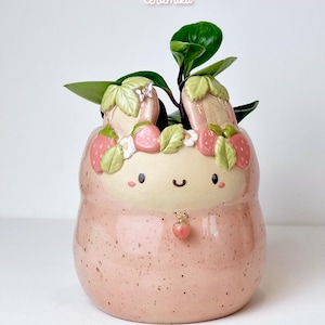 Peut inclure: Un pot de fleurs en céramique rose avec un design de fraise et de lapin. Le pot a une finition tachetée et un visage mignon. Le haut est décoré de fraises et de feuilles, et il contient une plante verte. Le texte "Ceramiku" et "ceramiku shop etsy com" sont visibles.