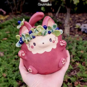 Può includere: Statuetta di coniglio in ceramica fatta a mano di colore rosa intenso, ornata da una corona di foglie e accenti di bacche blu. Il coniglio ha un viso e dettagli delle zampe color crema. Il logo "Ceramiku" è visibile.