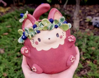 May include: A handcrafted ceramic bunny figurine in a deep rose color, adorned with a leafy crown and blue berry accents. The bunny has a cream-colored face and paw details. The logo "Ceramiku" is visible.