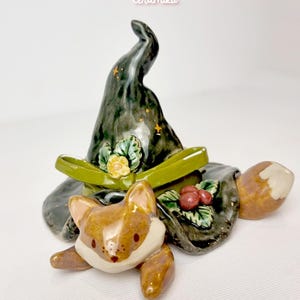 Op de afbeelding: Een keramieken vossenbeeldje met een heksenhoed. De hoed is donkergroen met gouden sterren en een groen lint. De vos is bruin en wit met een grillig ontwerp. Het beeldje heeft een decoratief bloemenaccent.