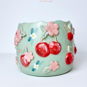 Pode incluir: Um vaso de cer&acirc;mica verde claro com uma borda ondulada, decorado com cerejas vermelhas, flores cor-de-rosa e pequenas flores brancas. O vaso tem os textos "Ceramiku" e "ceramikushop.etsy.com".