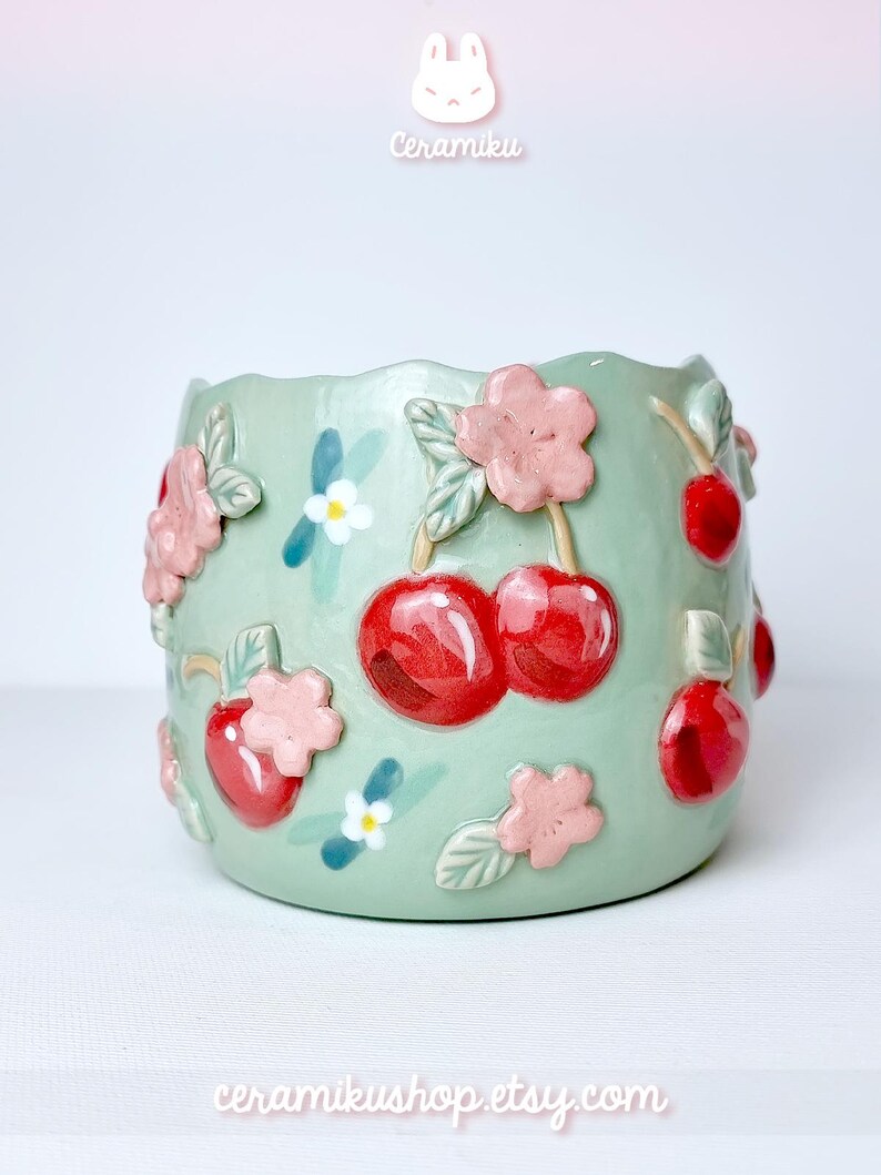 Pode incluir: Um vaso de cer&acirc;mica verde claro com uma borda ondulada, decorado com cerejas vermelhas, flores cor-de-rosa e pequenas flores brancas. O vaso tem os textos "Ceramiku" e "ceramikushop.etsy.com".