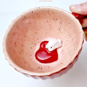 May include: A small, pink ceramic bowl with a scalloped edge. Inside, a red heart-shaped design holds a white bunny figurine. The bowl has a speckled texture and the text "ceramiku shop etsy.com" is visible.