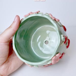 Pode incluir: Um vaso de cer&acirc;mica verde claro com detalhes florais rosa e vermelhos. O vaso tem formato redondo e um orif&iacute;cio de drenagem. Os textos "Ceramiku" e "ceramikusshop.etsy.com" s&atilde;o vis&iacute;veis.