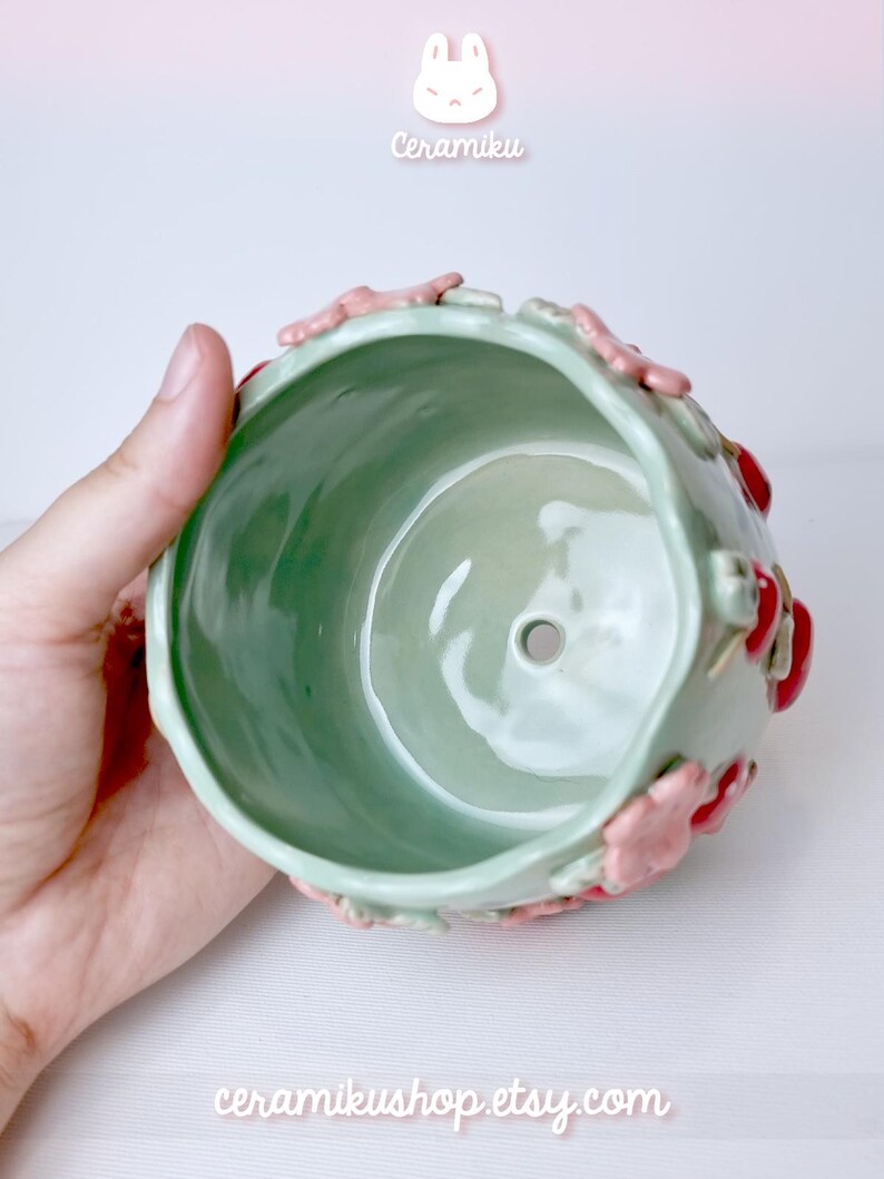 Pode incluir: Um vaso de cer&acirc;mica verde claro com detalhes florais rosa e vermelhos. O vaso tem formato redondo e um orif&iacute;cio de drenagem. Os textos "Ceramiku" e "ceramikusshop.etsy.com" s&atilde;o vis&iacute;veis.