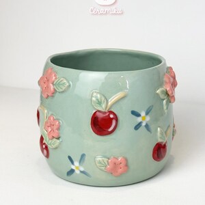 Puede incluir: Una taza de cerámica verde claro con forma redondeada. La taza está decorada con flores rosas en relieve, cerezas rojas y pequeños detalles florales azules y blancos. La marca "Ceramiku" está encima de la taza.
