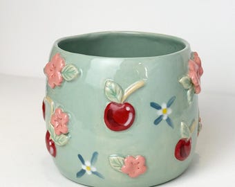 May include: A light green ceramic mug with a rounded shape. The mug is decorated with raised pink flowers, red cherries, and small blue and white floral accents. The brand name "Ceramiku" is above the mug.