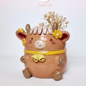 May include: A ceramic cow-shaped planter in shades of brown, with a yellow band and bell accents. The planter features a sad face, small horns, and yellow flower details. The planter is filled with dried flowers. The brand name "Ceramiku" is visible.