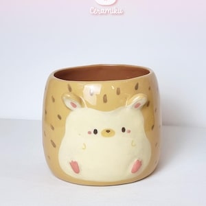 Op de afbeelding: Beige keramische mok met een hamsterontwerp. De mok heeft bruine stippen en een lichtbeige hamstergezicht met roze details. De binnenkant is bruin. De merknaam "Ceramiku" is bovenaan zichtbaar.