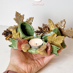 Op de afbeelding: Een keramische kandelaar met een herfstthema. Het heeft groene, bruine en gele bladeren, paddenstoelen en een groen lint. In het midden staat een brandende witte kaars. Handgemaakt.