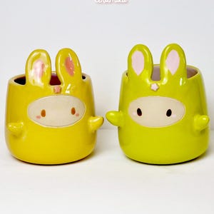 Peut inclure: Deux pots de fleurs en céramique en forme de lapin, un jaune et un vert, avec un visage blanc et des accents roses. Les pots ont de petits bras et des oreilles de lapin. Le texte "Ceramiku" et "ceramikusshop.etsy.com" sont visibles.