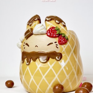 Op de afbeelding: Een keramische konijnvormige container met een geel wafelkegelpatroon. Het gezicht van het konijn is lichtbruin met chocoladedruppels, slagroom en rode aardbeienaccenten. De tekst "Ceramiku" en "ceramikusshop.etsy.com" zijn zichtbaar.