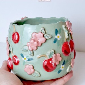 Pode incluir: Um vaso de cer&acirc;mica verde claro com uma borda ondulada, adornado com cerejas vermelhas em relevo, flores cor-de-rosa e pequenas flores brancas e azuis. Os textos "Ceramiku" e "ceramikushop.etsy.com" s&atilde;o vis&iacute;veis.
