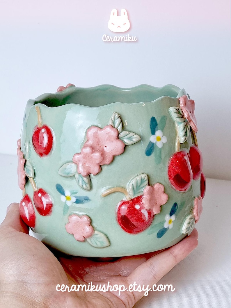 Pode incluir: Um vaso de cer&acirc;mica verde claro com uma borda ondulada, adornado com cerejas vermelhas em relevo, flores cor-de-rosa e pequenas flores brancas e azuis. Os textos "Ceramiku" e "ceramikushop.etsy.com" s&atilde;o vis&iacute;veis.