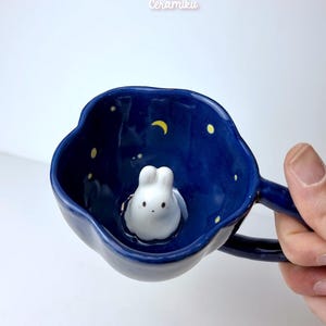Könnte beinhalten: Eine dunkelblaue Keramiktasse mit Henkel, mit einer weißen Hasenfigur im Inneren. Der Innenraum hat ein Nachthimmel-Design mit gelben Sternen und einem Mond. Die Tasse wird von einer Hand gehalten, mit dem Text "ceramikusshop.etsy.com" unten.