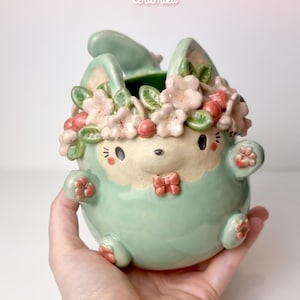 May include: A ceramic bunny planter in a light green hue, adorned with a floral crown of pink blossoms and red cherries. The bunny has a cream-colored face, a red bow, and small paws. The Etsy shop name "Ceramiku" is visible at the top.