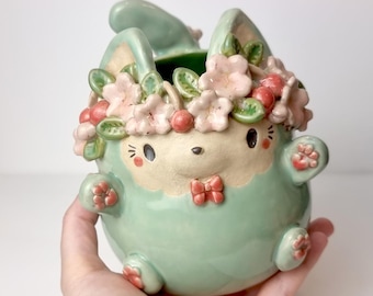 May include: A ceramic bunny planter in a light green hue, adorned with a floral crown of pink blossoms and red cherries. The bunny has a cream-colored face, a red bow, and small paws. The Etsy shop name "Ceramiku" is visible at the top.