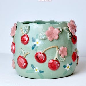 Pode incluir: Um vaso de cer&acirc;mica verde claro com uma borda ondulada, adornado com cerejas vermelhas, flores cor-de-rosa e flores brancas. O vaso tem o texto "Ceramiku" e "ceramikusshop.etsy.com".