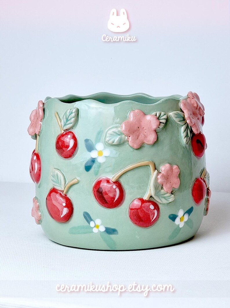 Pode incluir: Um vaso de cer&acirc;mica verde claro com uma borda ondulada, adornado com cerejas vermelhas, flores cor-de-rosa e flores brancas. O vaso tem o texto "Ceramiku" e "ceramikusshop.etsy.com".
