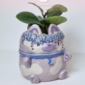 Puede incluir: Una maceta de cerámica de color morado claro con forma de gato y manchas blancas, que lleva un collar azul con un pequeño cascabel y una corona de flores azules. Una planta verde crece en la parte superior. Aparece el texto Ceramiku y Ceramikushop.etsy.com.