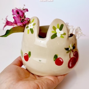 May include: A beige ceramic bunny-shaped planter with red cherry and white flower designs. The planter holds pink and white flowers. The text "Ceramiku" and "ceramikusshop.etsy.com" are visible.