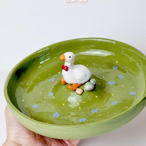 Può includere: Un piatto in ceramica verde con una statuina di oca bianca con un papillon rosso. Il piatto presenta decorazioni floreali blu e una piccola statuina di oca. Il testo "ceramiku" e "ceramikusshop.etsy.com" sono visibili.