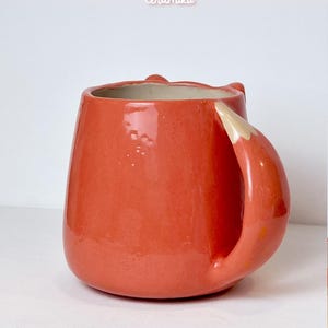 K&ouml;nnte beinhalten: Eine Keramiktasse in leuchtendem Orange, geformt wie ein Fuchs. Der Griff ist so gestaltet, dass er einem Fuchsschwanz &auml;hnelt, mit einer wei&szlig;en Spitze. Die Tasse tr&auml;gt die Texte "Ceramiku" und "ceramikushop.etsy.com".