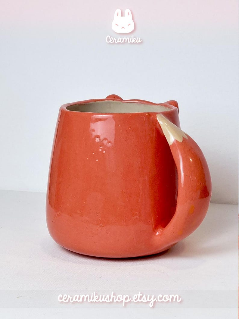 K&ouml;nnte beinhalten: Eine Keramiktasse in leuchtendem Orange, geformt wie ein Fuchs. Der Griff ist so gestaltet, dass er einem Fuchsschwanz &auml;hnelt, mit einer wei&szlig;en Spitze. Die Tasse tr&auml;gt die Texte "Ceramiku" und "ceramikushop.etsy.com".