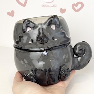 Puede incluir: Un recipiente de cerámica negra brillante con forma de gato con un colgante de estrella en su collar. El recipiente tiene un cuerpo redondeado, una cola rizada y pequeñas patas. Decorado con detalles en rosa y la palabra "Ceramiku".