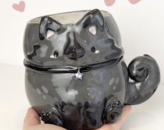 Op de afbeelding: Een glanzende zwarte keramische katvormige container met een sterhanger aan de halsband. De container heeft een afgerond lichaam, een gekrulde staart en kleine pootjes. Versierd met roze accenten en het woord "Ceramiku".
