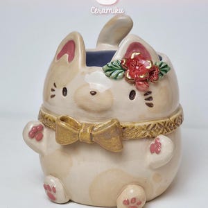 Puede incluir: Un recipiente de cerámica con forma de gato en crema y beige. Tiene un lazo dorado, una flor roja y el texto "CeramikuShop.etsy.com". El gato tiene detalles rosas en las orejas y las patas.