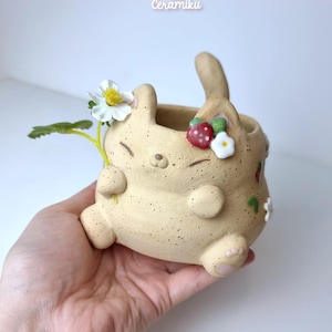 Puede incluir: Maceta de cerámica beige con forma de conejo, adornada con fresas y flores. La maceta tiene una textura moteada y sostiene una flor. El sitio web ceramikushop.etsy.com es visible en la parte inferior.