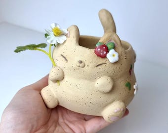 Op de afbeelding: Een beige keramische konijnenplantenbak versierd met aardbeien en bloemen. De pot heeft een gespikkelde textuur en houdt een bloem vast. De website ceramikushop.etsy.com is zichtbaar onderaan.