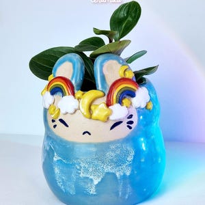 Op de afbeelding: Een blauwe keramische plantenbak in de vorm van een konijn, versierd met regenbogen, wolken en sterren. De plantenbak heeft een glanzende afwerking en bevat groene bladplanten. De teksten "Ceramiku" en "ceramikusshop.etsy.com" zijn zichtbaar.