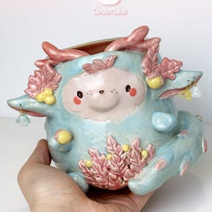 May include: A whimsical, light blue ceramic planter with a cartoonish face and pink accents. The planter features a smiling face, decorative pink leaf-like elements, and small dangling charms. The word "Ceramiku" is visible in the upper center.