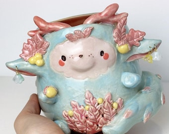 May include: A whimsical, light blue ceramic planter with a cartoonish face and pink accents. The planter features a smiling face, decorative pink leaf-like elements, and small dangling charms. The word "Ceramiku" is visible in the upper center.