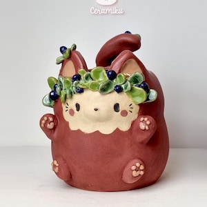 May include: A ceramic planter in the shape of a cartoon bunny, in a reddish-brown hue. The bunny has a white face, a green and blue floral crown, and paw-shaped details. The brand name "Ceramiku" is above the planter.