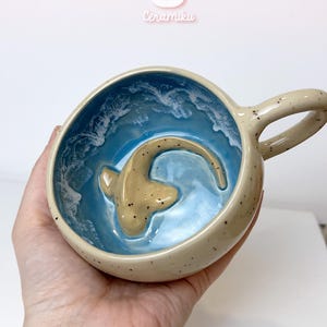 May include: A handmade ceramic mug with a beige exterior and a blue interior, featuring a beige shark design. The mug has a handle and is speckled with dark brown spots. The word "Ceramiku" is above the mug.