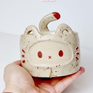 Op de afbeelding: Een gespikkelde keramische katvormige plantenbak met een rood gekleurde staart en rode gezichtskenmerken. De plantenbak heeft een kleine gouden bedel die aan de kraag hangt. De tekst "Ceramiku" en "ceramikusshop.etsy.com" zijn zichtbaar.