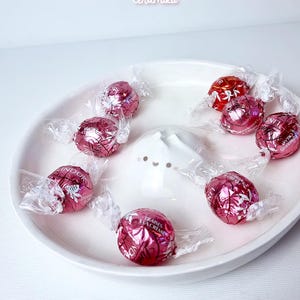 May include: A white ceramic dish with a raised, cat-shaped design in the centre. The dish is filled with pink and red chocolate candies wrapped in clear plastic.