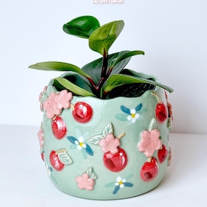Pode incluir: Um vaso de cer&acirc;mica verde claro com um design de cerejas e flores, contendo uma planta verde. O vaso tem cerejas vermelhas em relevo e flores cor-de-rosa. O texto "Ceramiku" e "ceramikusshop.etsy.com" s&atilde;o vis&iacute;veis.