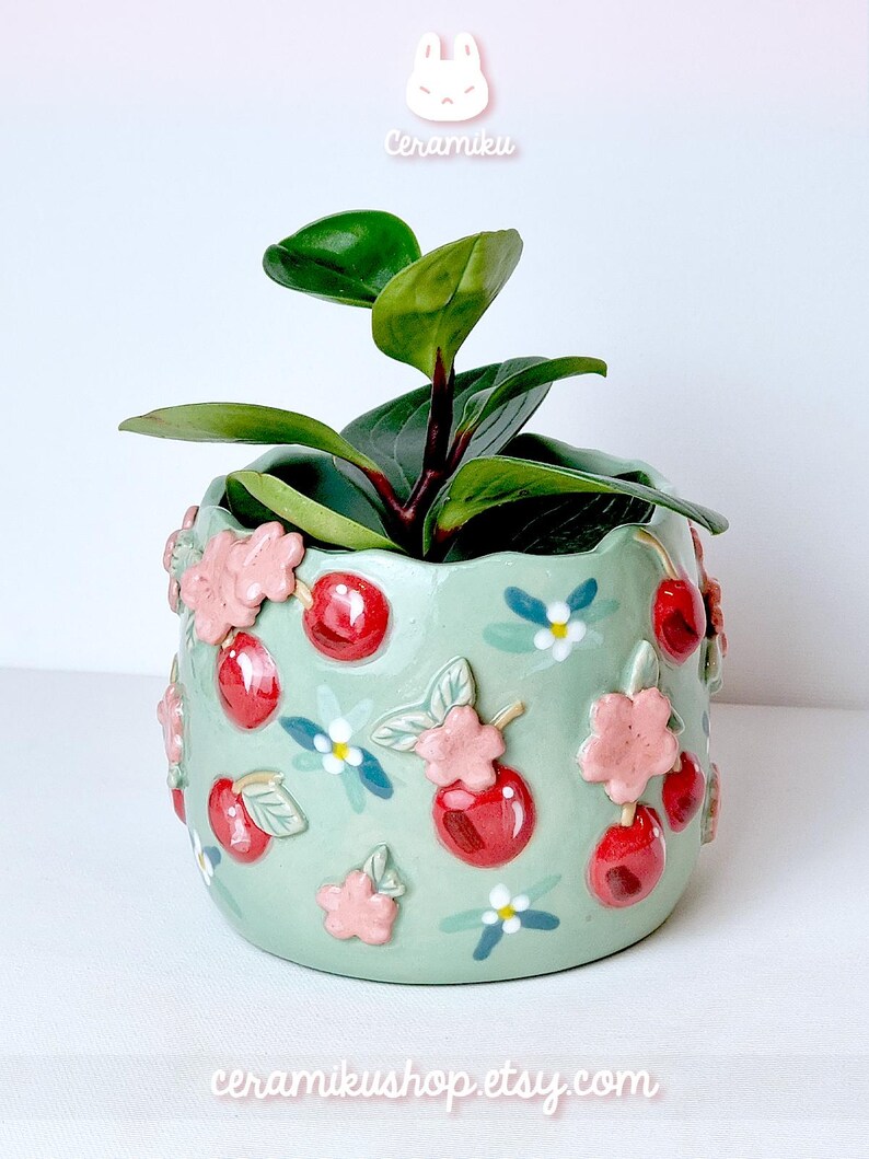 Pode incluir: Um vaso de cer&acirc;mica verde claro com um design de cerejas e flores, contendo uma planta verde. O vaso tem cerejas vermelhas em relevo e flores cor-de-rosa. O texto "Ceramiku" e "ceramikusshop.etsy.com" s&atilde;o vis&iacute;veis.