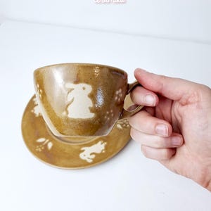 Könnte beinhalten: Ein braunes Keramik-Tassen- und Untertassen-Set mit Hasenmuster. Die Tasse hat einen Henkel und eine weiße Hasensilhouette an der Seite. Die Untertasse hat ebenfalls Hasenmuster. Der Text "Ceramiku" und "ceramikusshop.etsy.com" sind sichtbar.