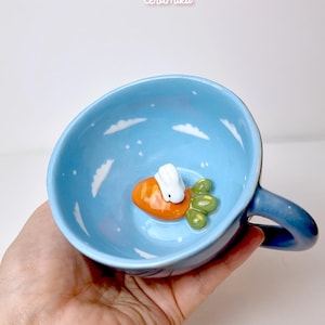 May include: A light blue ceramic mug with a whimsical design. Inside the mug, a white bunny sits on an orange carrot, surrounded by green leaves. The interior features white cloud and star details. The mug has a handle and the text "ceramiku shop.etsy.com".