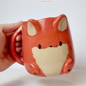 Peut inclure: Mug en céramique en forme de renard, de couleur orange et crème. Le mug présente des oreilles, des pattes et un visage de renard. Le texte "Ceramiku" et "ceramikusshop.etsy.com" sont visibles.
