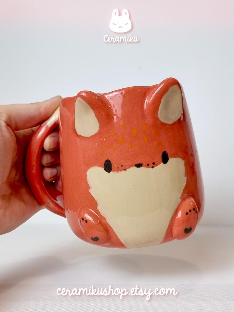 K&ouml;nnte beinhalten: Keramikbecher in Fuchsform mit orangefarbenen und cremefarbenen Farbt&ouml;nen. Der Becher hat Fuchsohren, Pfoten und ein Gesicht. Der Text "Ceramiku" und "ceramikusshop.etsy.com" sind sichtbar.