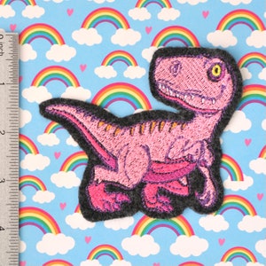 Pode incluir: Patch de dinossauro bordado rosa com um suporte de feltro cinza. O dinossauro está em uma pose de corrida com a boca aberta e um olho amarelo.