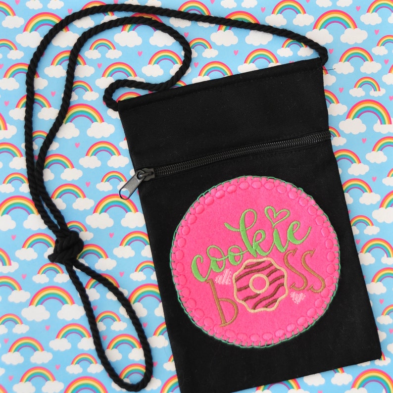 May include: Black canvas zippered pouch with an adjustable strap. The pouch has a pink circle with the text "cookie boss" and a donut graphic in the center.