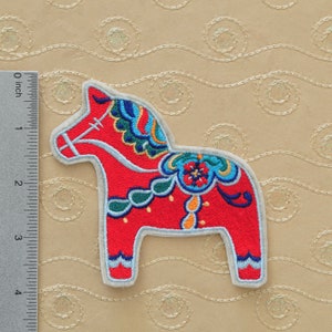 May include: A red and blue embroidered Dala horse, a traditional Swedish folk art wooden toy, with a white outline. The horse is about 4 inches tall.
